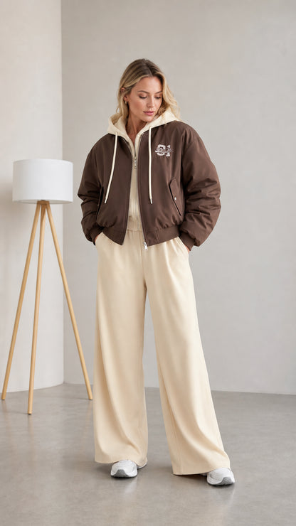 Front full view of woman wearing brown layered hooded bomber jacket with cream wide-leg pants