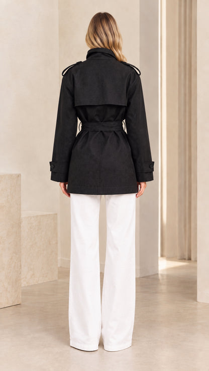 Back view of model wearing black short trench coat showing storm flap detail and tie belt