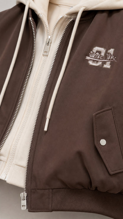 Close-up detail of brown bomber jacket showing double zipper, hood drawstring, and chest embroidery