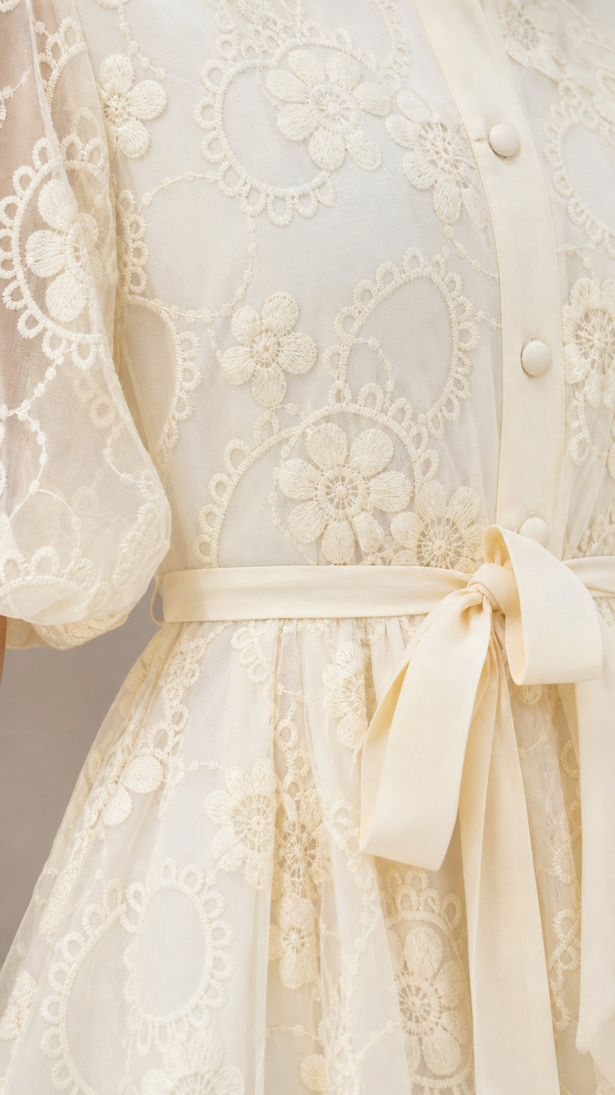 Close-up detail of cream embroidered floral lace, covered buttons, and waist sash