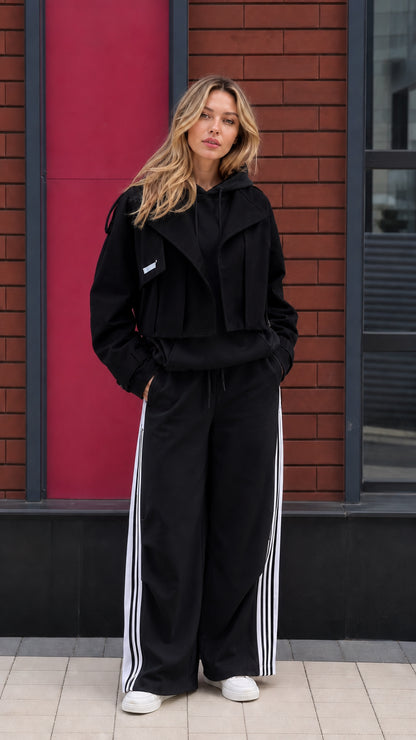 Model wearing black hoodie and black wide leg sweatpants set with white side stripes and cropped jacket