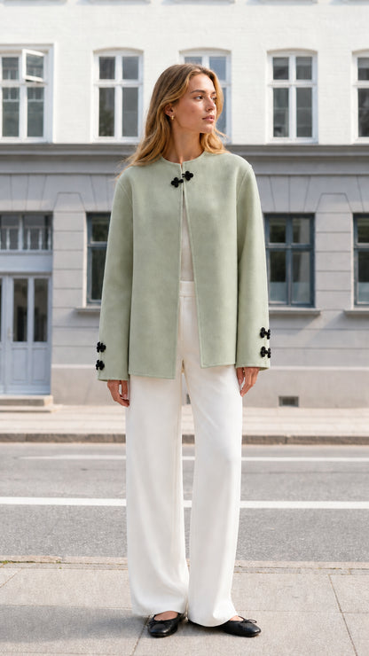 Front full view of woman wearing sage green collarless knot clasp jacket with white trousers
