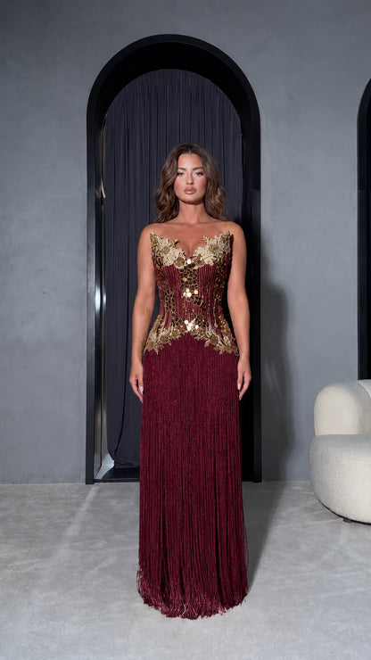Front view of model wearing Burgundy strapless evening dress with gold embellished bodice and wine red fringe